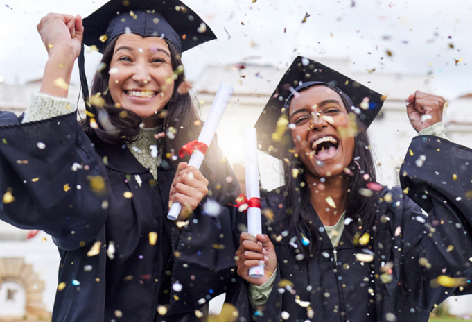 Two celebrating females in caps and gowns surrounded by confetti in the air