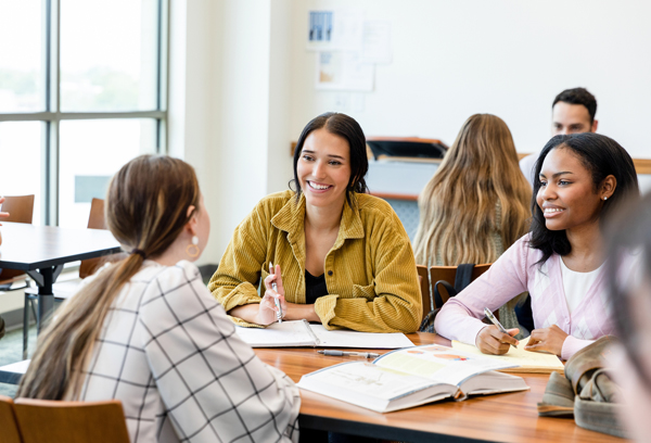 Taking a break from their assignment, the two young adult female friends smile as they listen to the unrecognizable young adult woman seated across from them