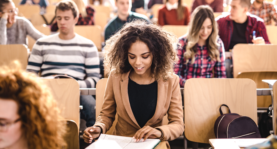 Happy female student attending a class with her classmates at lecture hall and reading a textbook