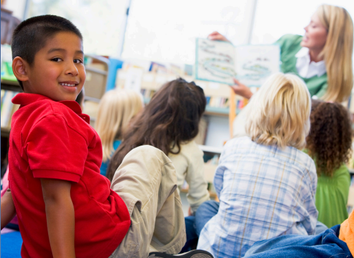 The back of a reading group of children in the background with a smiling young boy in the foreground turning around to face the camera